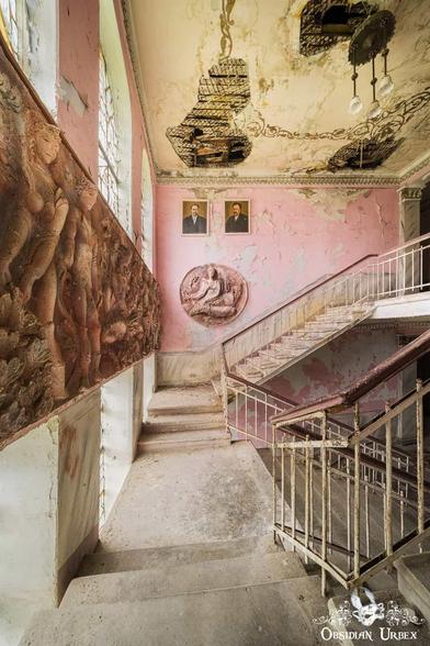 An abandoned pink staircase in a Soviet-era building shows decay, flaking paint, and crumbling ceilings.